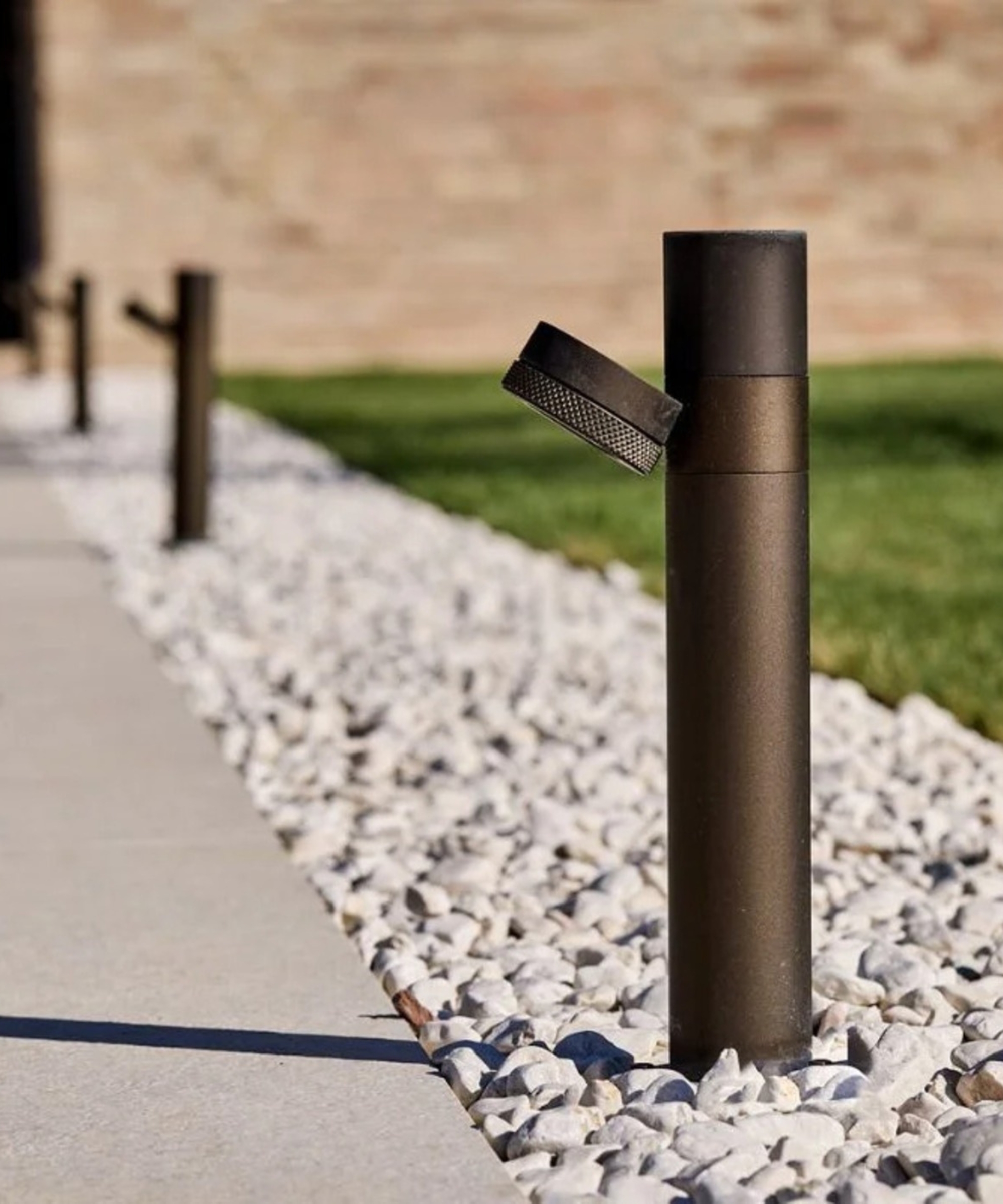 Bronze bollard light with an angled cap, set in white gravel between a paved path and lawn.