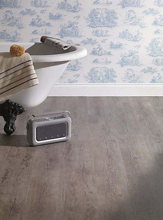 Wickes Caspian Grey Oak Luxury Vinyl Flooring in a bathroom with half a freestanding bathtub visible, with a olden style radio on the floor and a blue patterned wallpaper