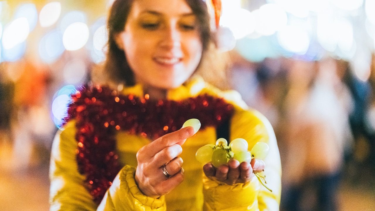 A woman in a yellow coat with red tinsel around her neck eats grapes outside during New Year&#039;s Eve festivities in Spain