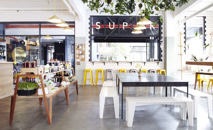 Inside Superette there are golden lights hanging from ceiling over black table and white benches. Yellow and gray stools are standing on the side.