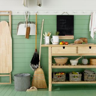 Utility room and kitchen, green and white tongue and groove panelling, hanging laundry dryer rack, peg rail on pale wood kitchen unit, straw broom, feather duster, hanging dustpan and brush, basket fruit storage with apples, blackboard notice board