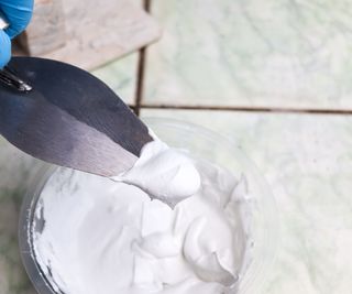 Someone using a metal trowel to mix white til grout in a clear plastic pot.