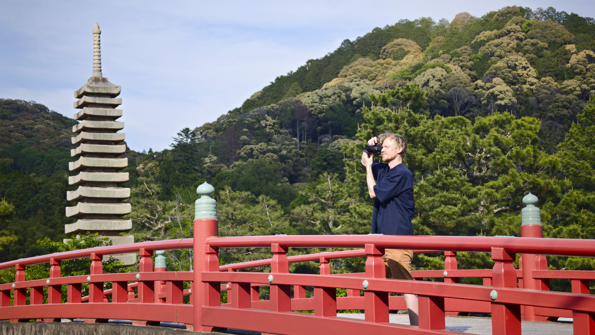 Man holding the Panasonic Lumix G1 mirrorless camera in a rural Japan setting, stood by a red bridge