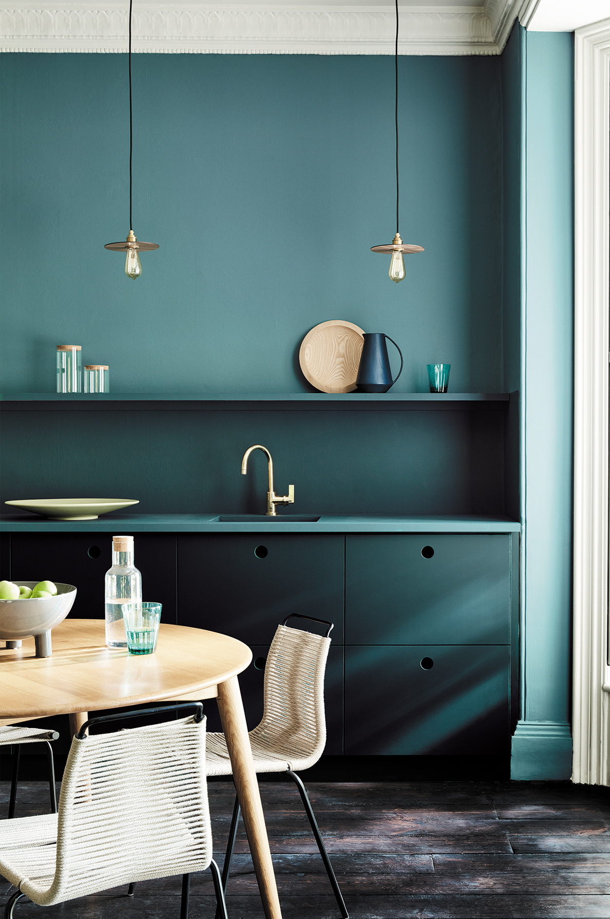 Modern kitchen with teal walls and dark green cabinets, a round wooden dining table, and woven chairs.
