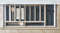 A plan view of an open kitchen drawer with wooden dividers and a navy liner housing black cutlery and wooden spoons