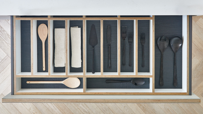 A plan view of an open kitchen drawer with wooden dividers and a navy liner housing black cutlery and wooden spoons