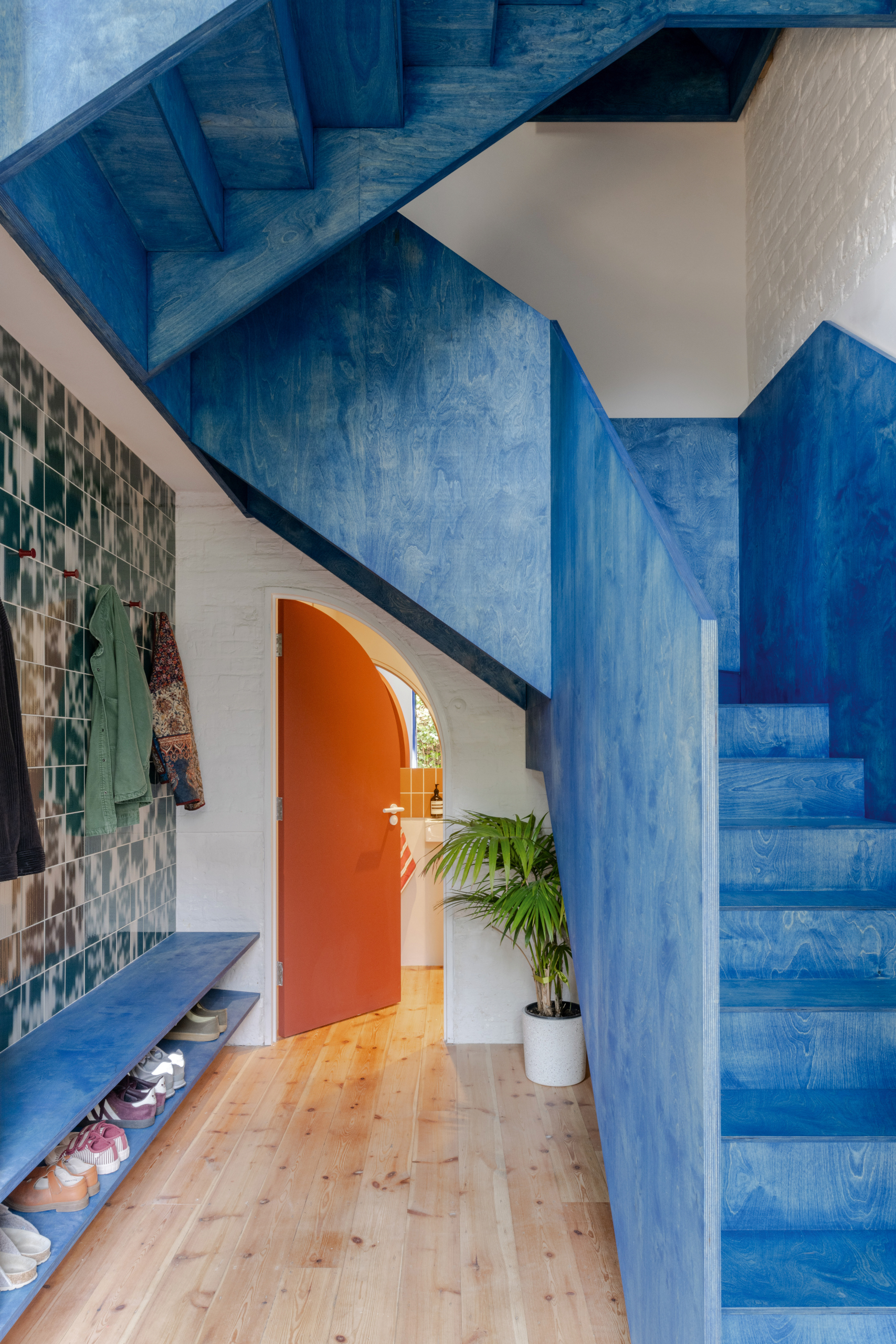 Hallway with angular enclosed timber staircase stained in bright blue, with a semi-circular orange door at the end of the hallway and a wall of tiles to the left.