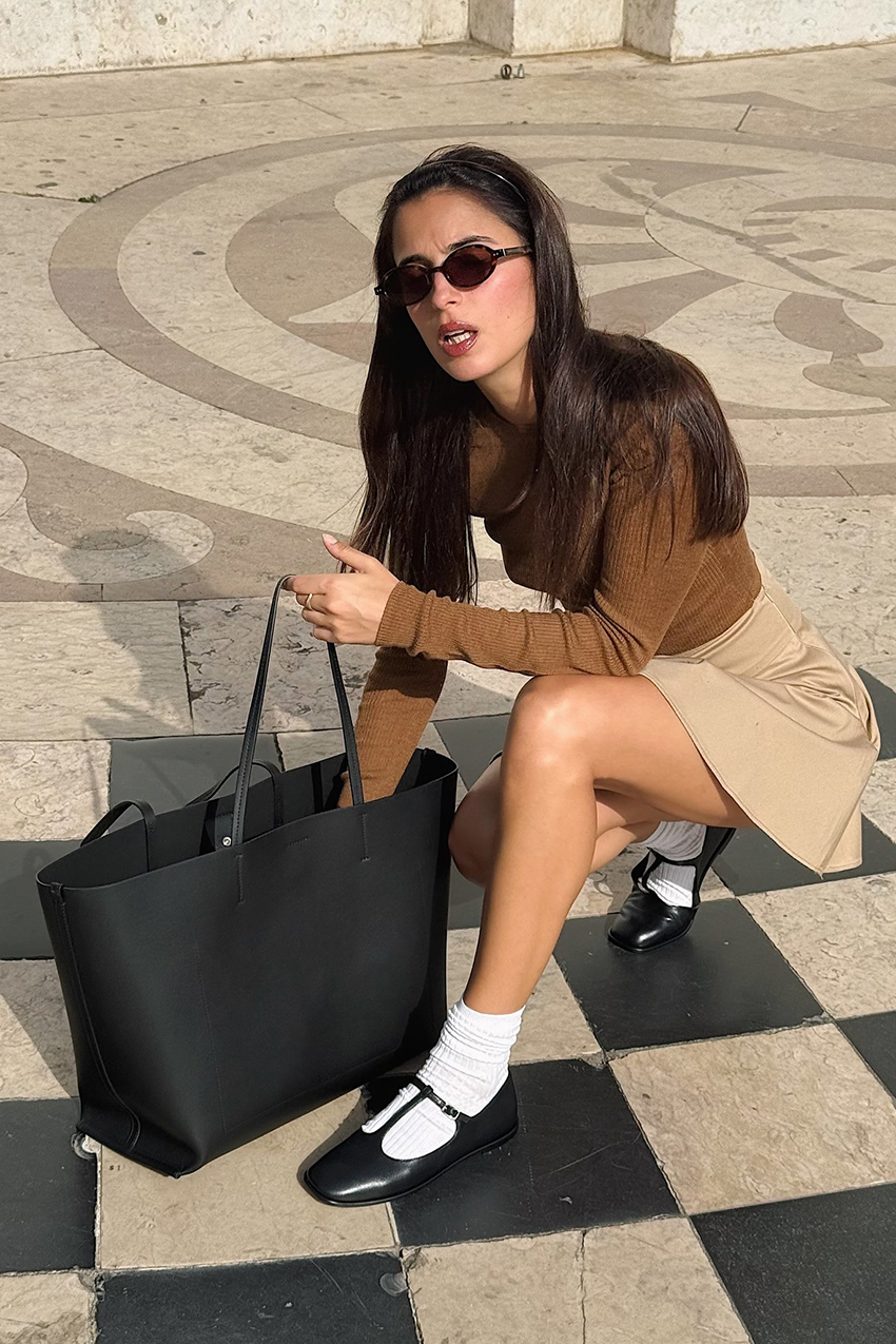 a woman wearing a brown shirt, tan skirt, black t-strap flats, white socks, and a black bag