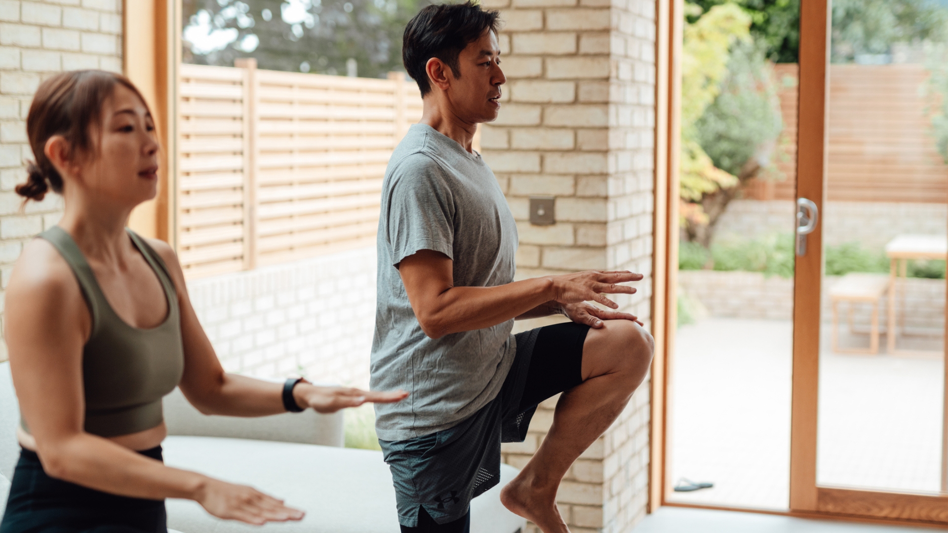 woman and a man marching on the spot with high knees sideways to the camera indoors with patio doors behind them. 