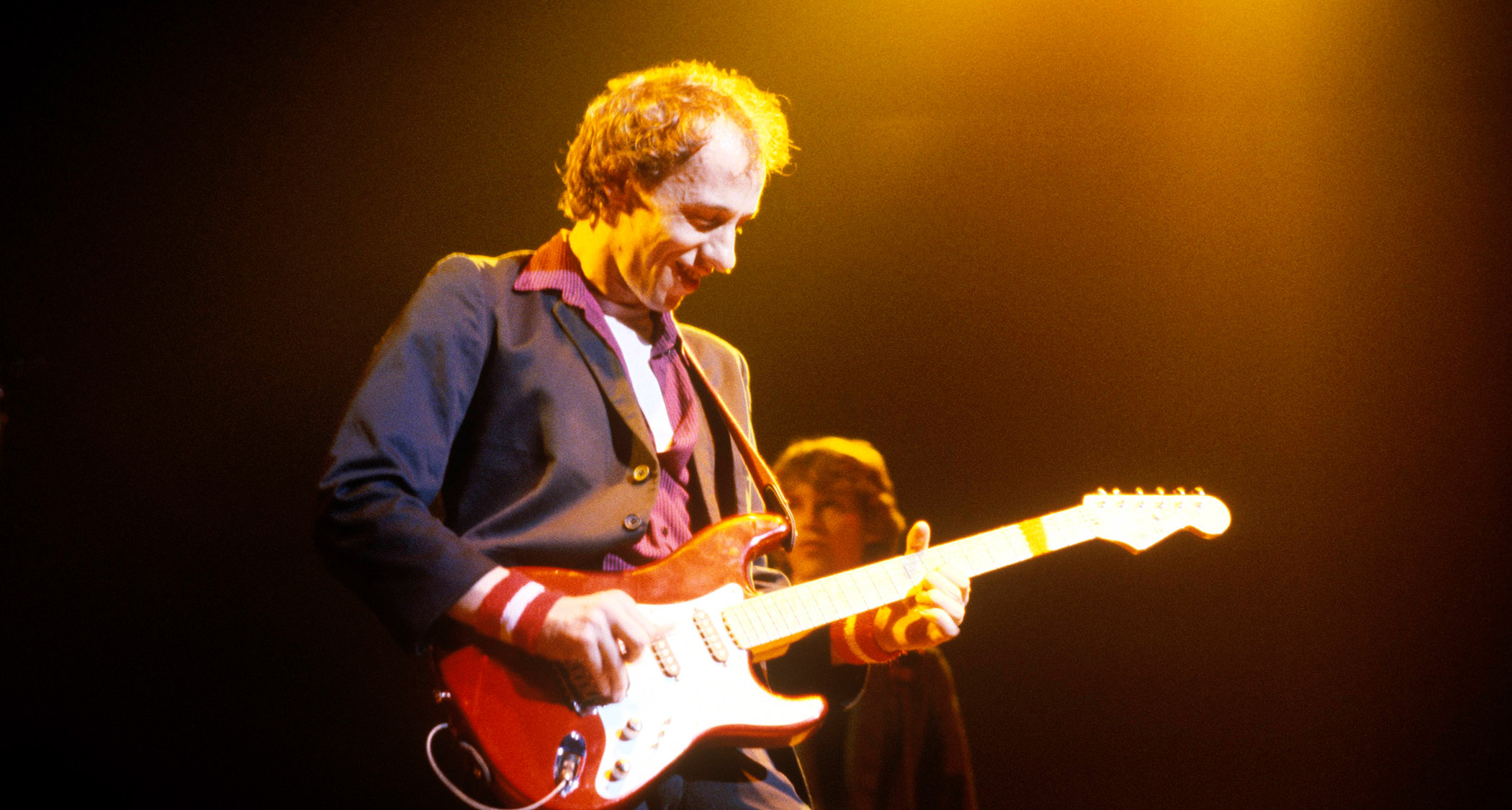 Mark Knopfler of Dire Straights performs onstage in 1985 with his red Fender Stratocaster. He wears a dark suit and a burgundy shirt with an open collar.