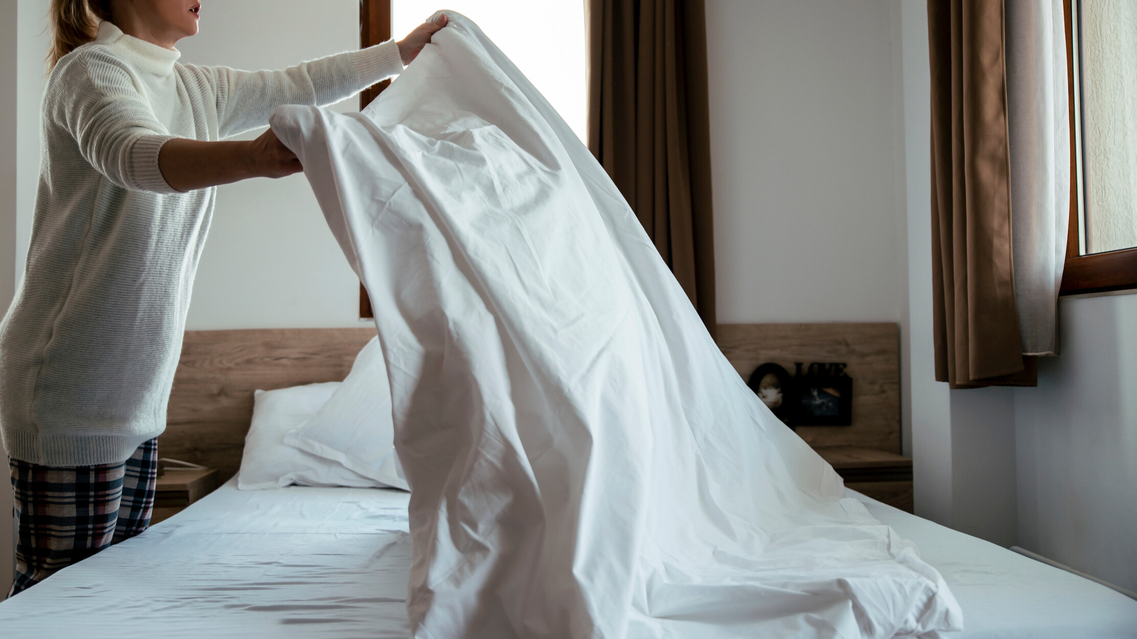 A woman making the bed in a using white bedding in a room