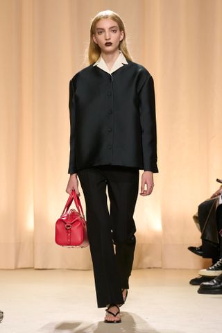 Bally model wearing a black suit with a yellow shirt underneath, a red bag, and flip flops.