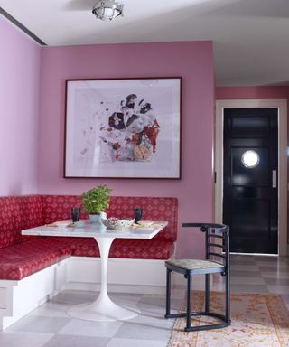Breakfast nook painted light pink with red patterned seating cushions o the banquette seating. A white square table topped with a plant sits in the middle while a large abstract artwork hangs on the wall