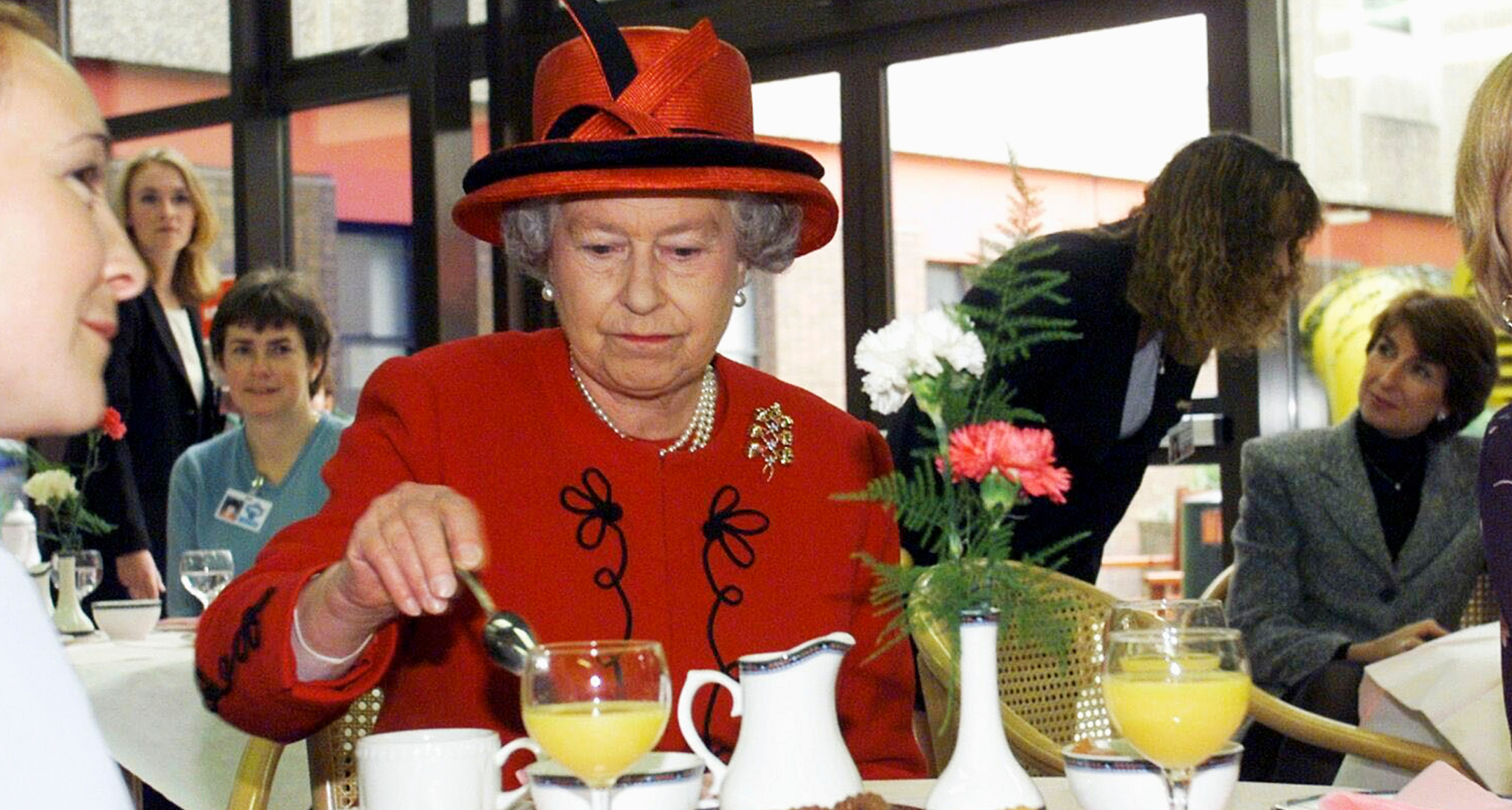 Queen Elizabeth wearing a red coat and hat stirring tea