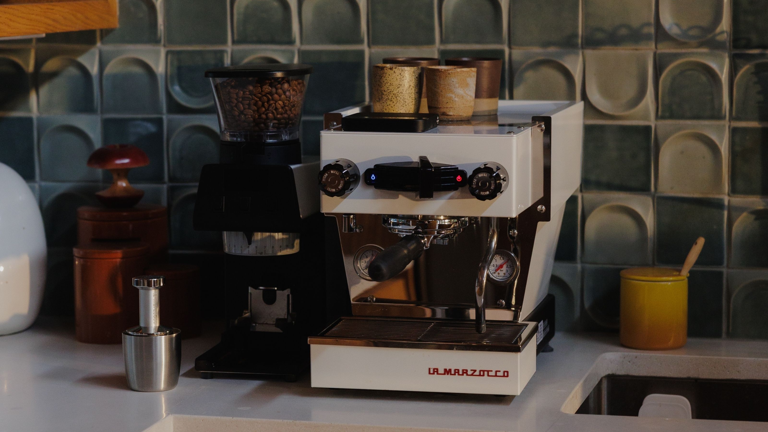 A white La Marzocco coffee machine next to a black coffee grinder on a white kitchen counter with blue backsplash.