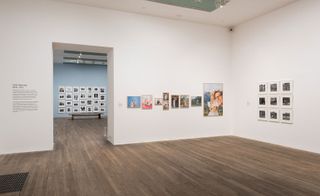 Wooden floor, white walls, white ceiling with lighting, picture gallery, black grate on the floor to the left, open doorway with view of the next room, pale blue walls and picture gallery
