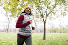 Woman running through park wearing winter workout clothes, holding water bottle and listening to music with wired headphones, smiling