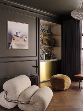 Image of a dark living room with a gold and marble built-in bookshelf. There is a textured white accent chair and an ochre yellow pouf on the floor.