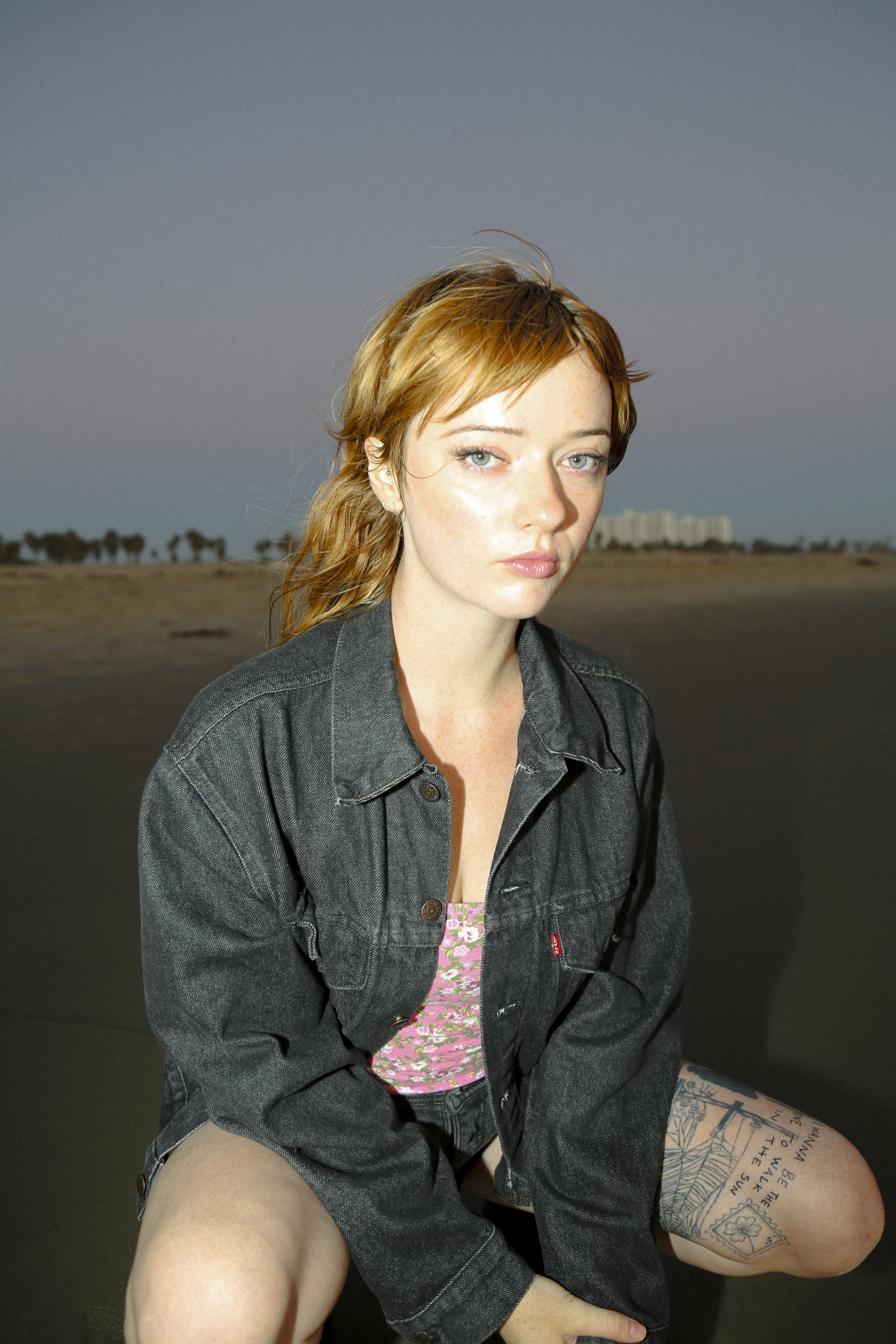 actress lindsey normington poses on a beach at sunset wearing a black jean jacket printed pink top and black denim shorts