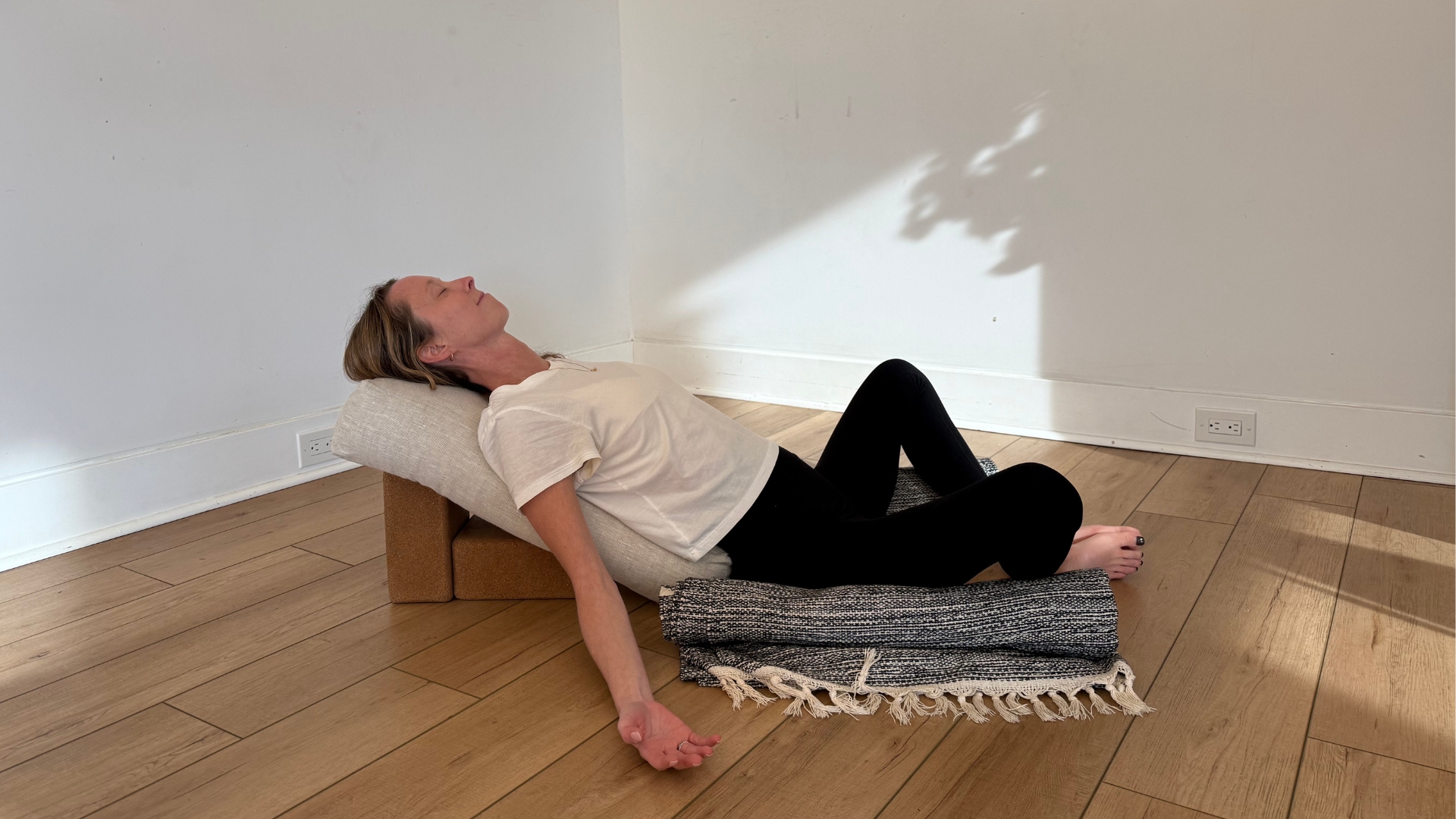 woman in reclined bound angle pose on a bolster and wooden floor with white walls behind her