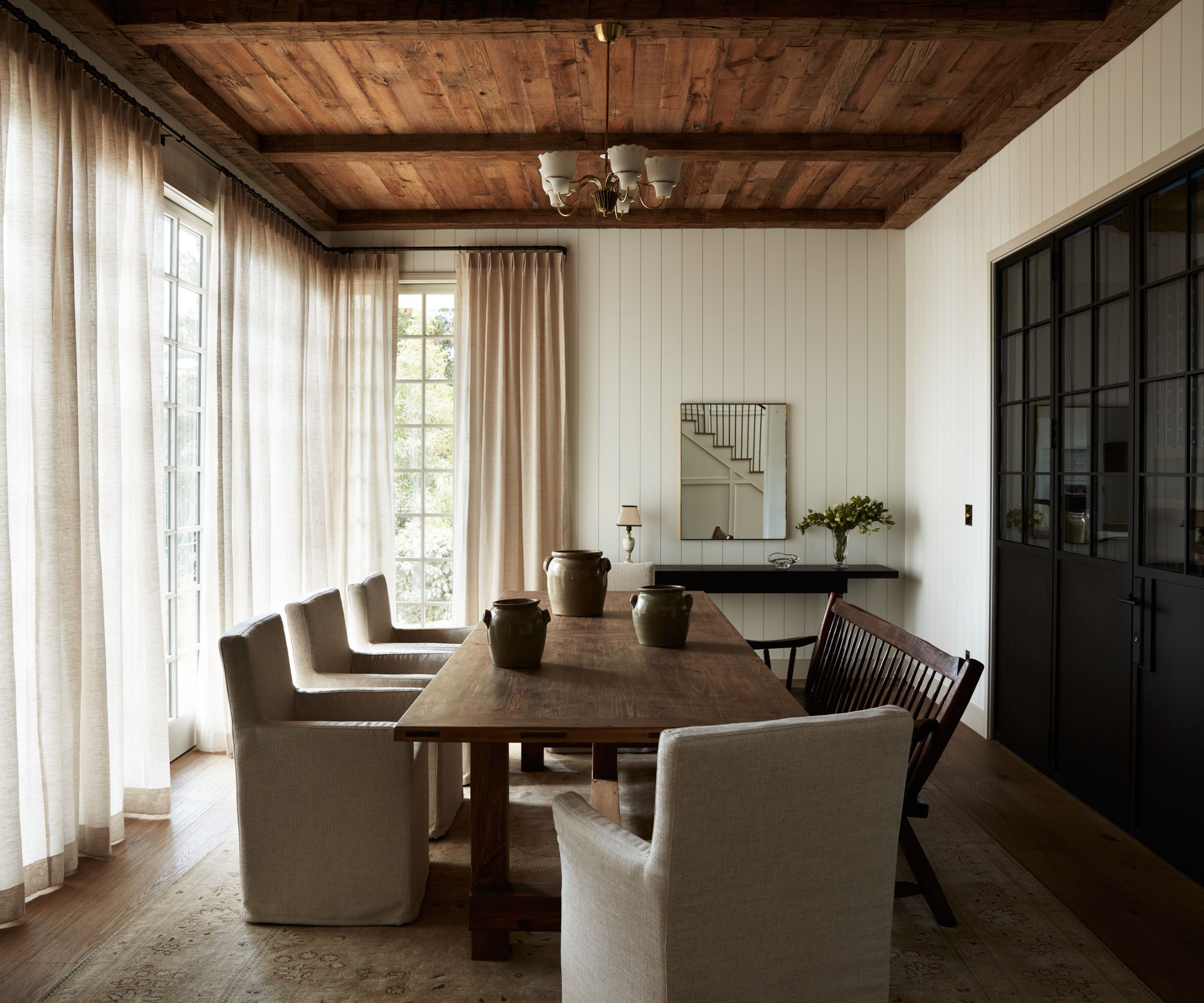 Neutral dining room with white panelled walls, long dark wood dining table with upholstered chairs and bench, sheer curtains on windows, black cabinetry