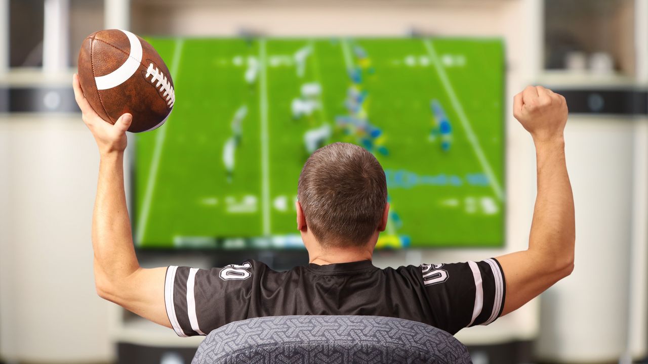 A man holding a football has his hands in the air to celebrate while watching a game on TV.