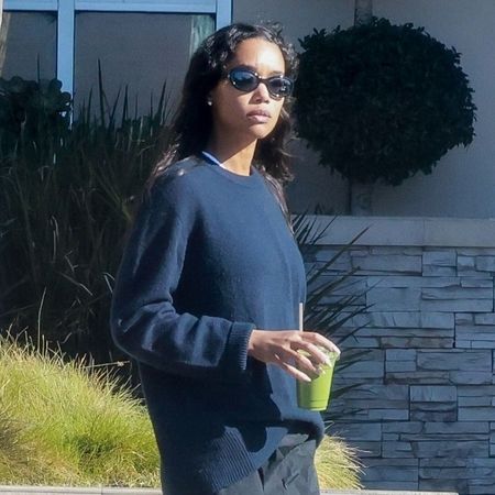 Laura Harrier wears grey trainers with black trousers and a navy jumper.