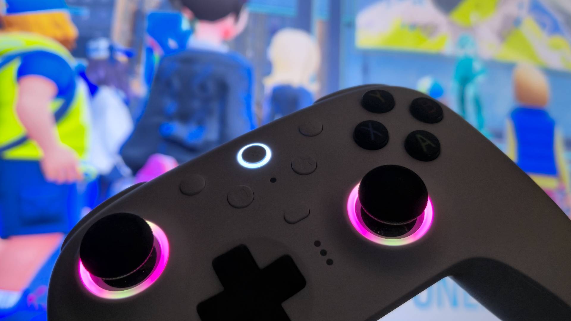 Photo showing a white controller being held up to a TV. The sticks of the controller are glowing pink and yellow.