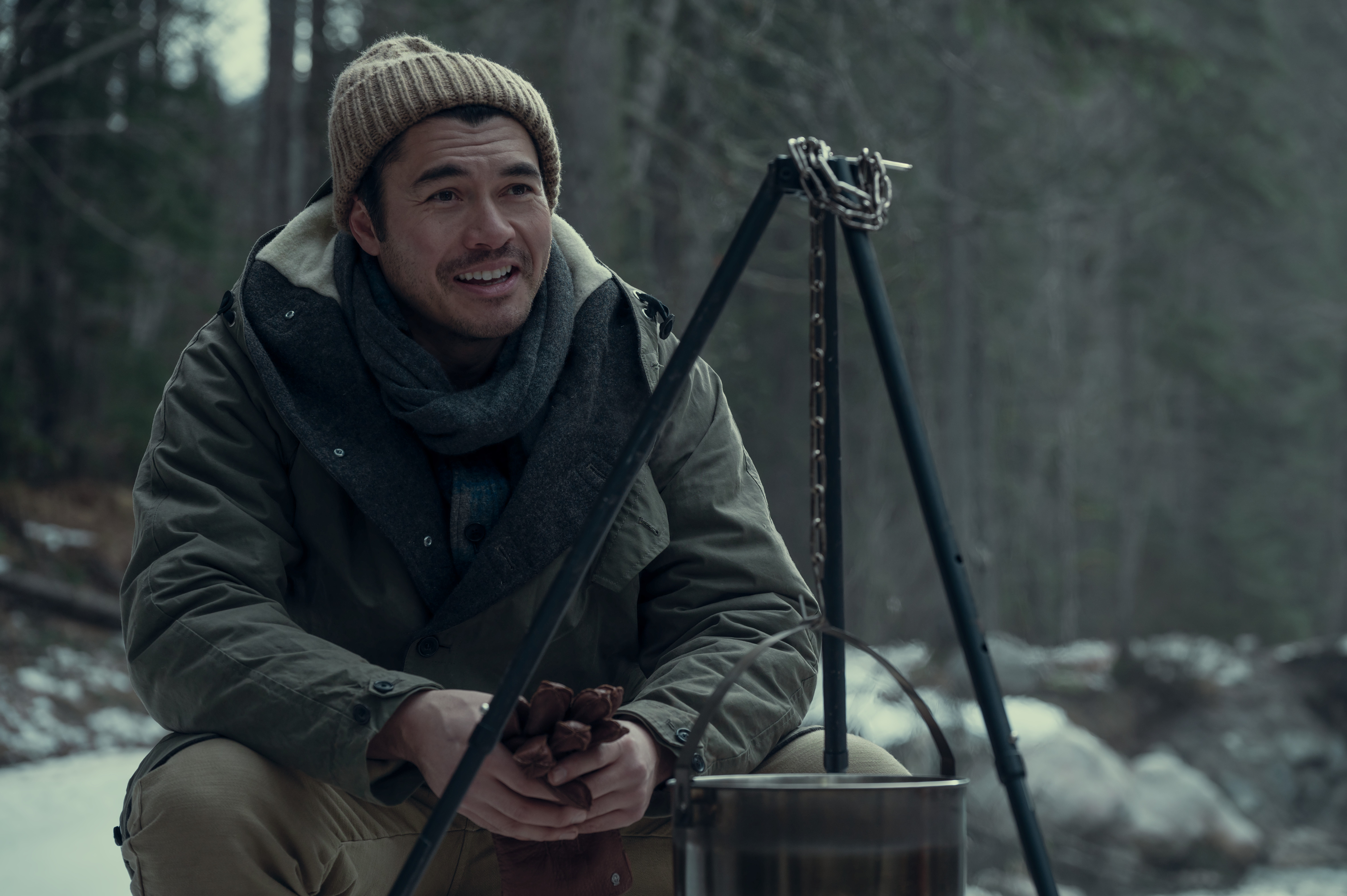 henry golding as peter wearing a beanie and coat sitting by a snowy stream in nine perfect strangers season 2