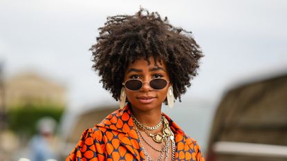 Sarah Monteil wears black sunglasses, oversized silver earrings, silver large chain necklaces, a navy blue with orange print pattern oversized silk shirt, high waist matching navy blue with orange print pattern large pants, outside the Thom Browne show, during Paris Fashion Week - Menswear Spring/Summer 2023, on June 26, 2022 in Paris, France. (