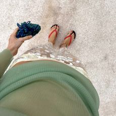 style influencer Courtney Grow poses on a sandy beach wearing red The Row jelly flip-flop sandals, a shell anklet, a white sheer embroidered skirt with sequins, a knitted clutch, and sheer ribbed green top