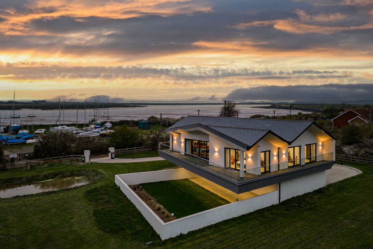 The house at night is lit by wall lighting around the balcony and the home looks out over the sea and boating dockyards