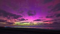 a beach scene at night where the silhouettes of people stand on the beach gazing up at the partly cloudy sky that is awash with pinks, purples, yellows and greens.