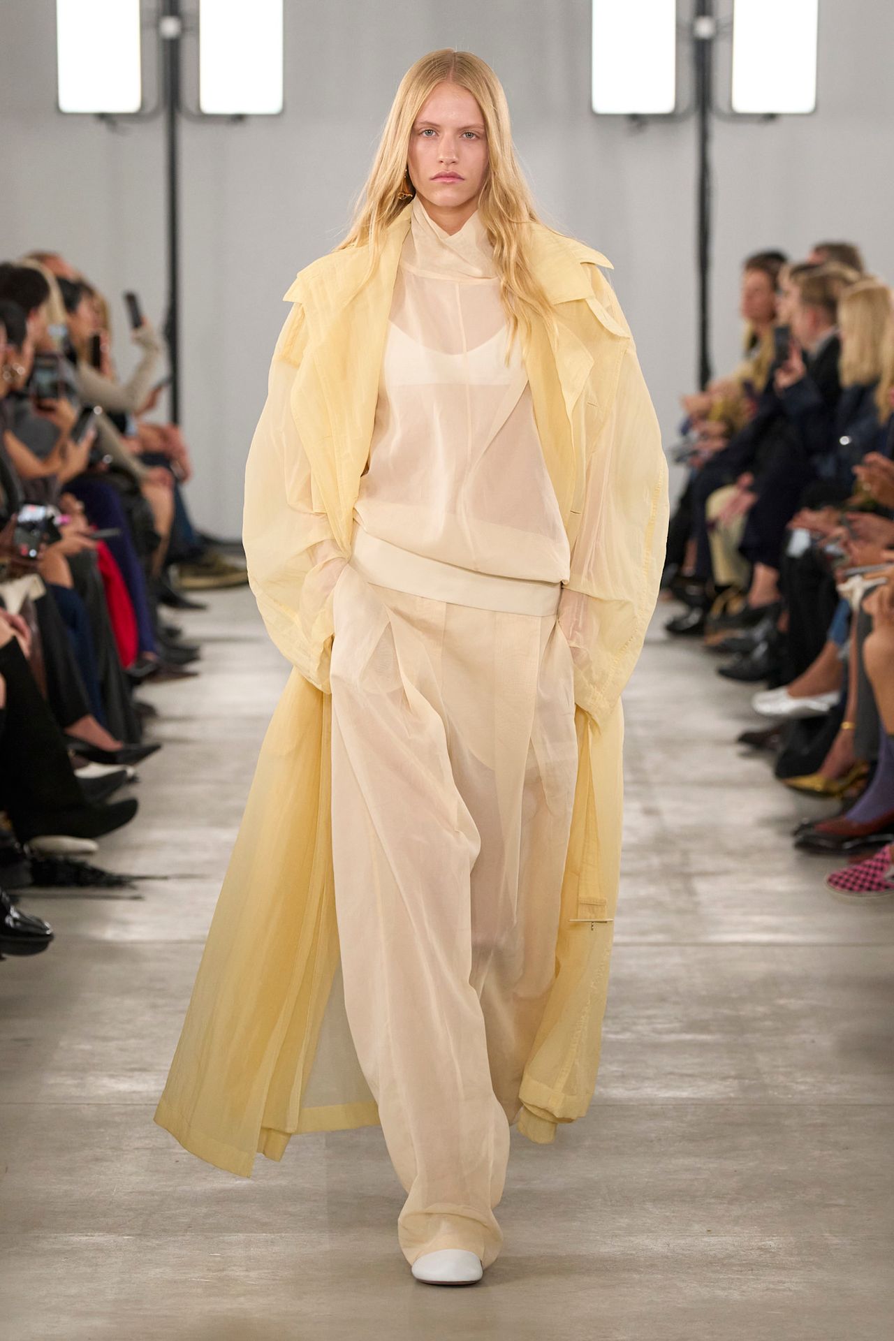 Layering staples for spring 2026 is shown in a photo from Sportmax's runway show, with a model walking while wearing a butter yellow sheer organza trench layered on top of a sheer high-neck white blouse and white bralette styled with matching wide-leg organza trousers and white ballet flats