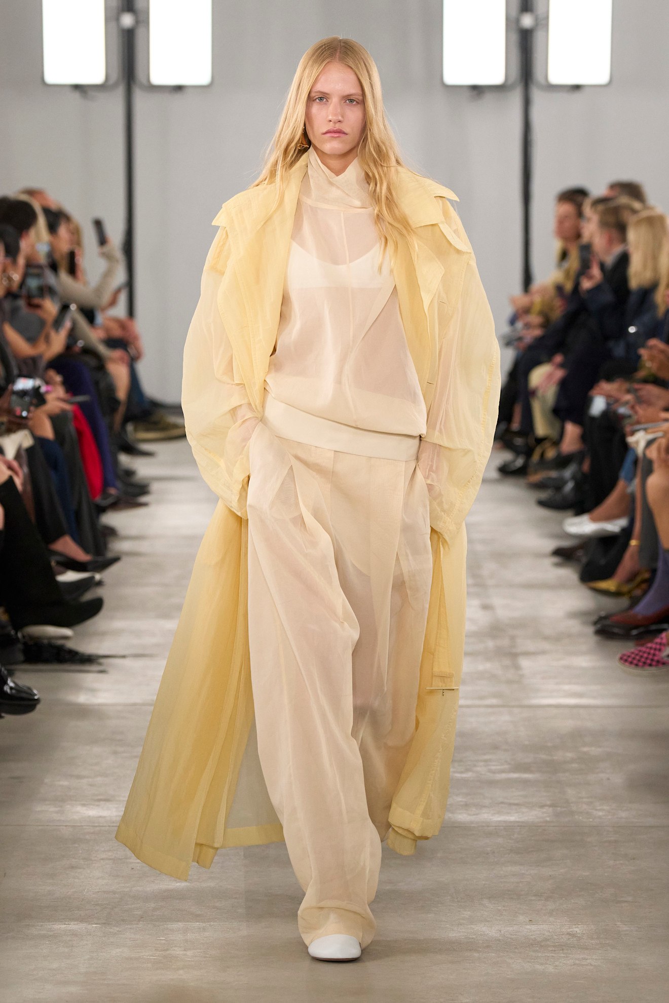 Layering staples for spring 2026 is shown in a photo from Sportmax's runway show, with a model walking while wearing a butter yellow sheer organza trench layered on top of a sheer high-neck white blouse and white bralette styled with matching wide-leg organza trousers and white ballet flats 