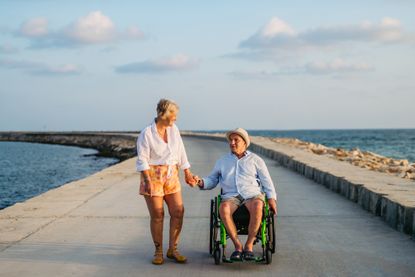 A woman in shorts and a man on a wheelchair holding hands on a boardwalk surrounded by water.