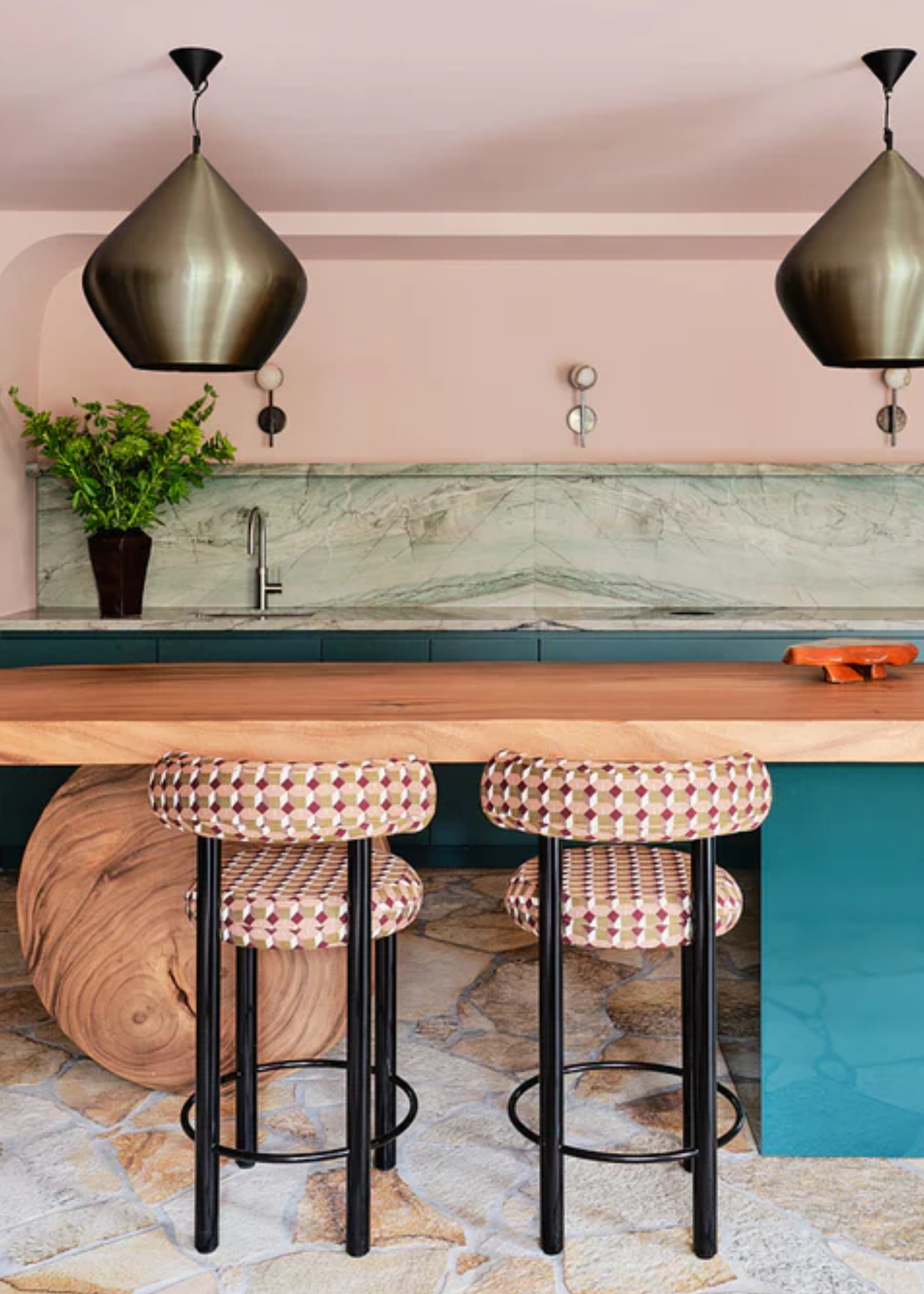 A pale pink kitchen with a green stone backsplash, a blue-green base, an island with a wood countertop and a round wood foot alongside a blue-green leg by a pair of patterned kitchen stools below brushed silver lights