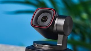 a small PTZ webcam with 4K resolution and a magnetic attachment clip is photographed in front of a blue background and a green plant