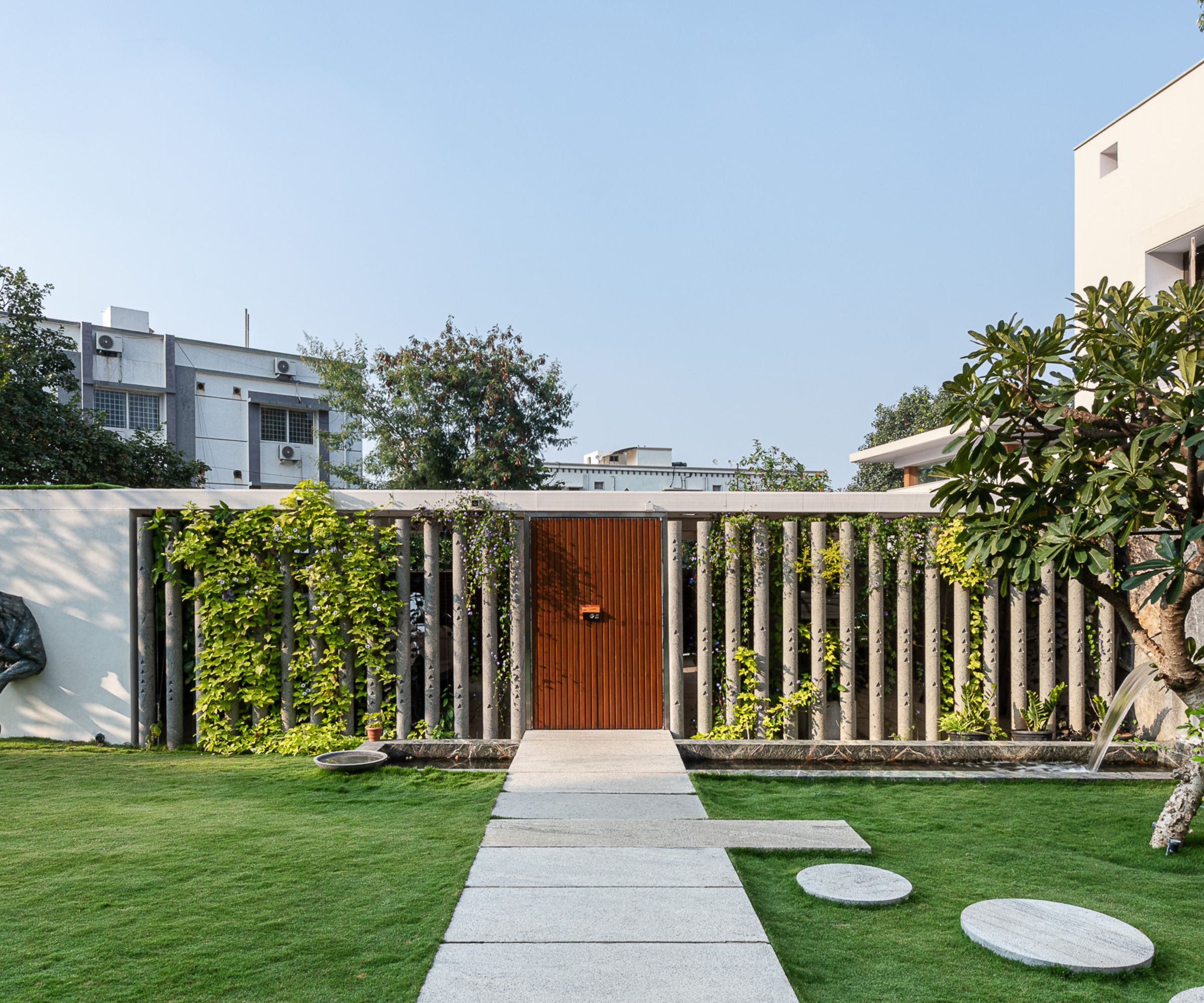 Wooden door in slatted concrete screen wall covered in climbing vines, stone path across a lawn.