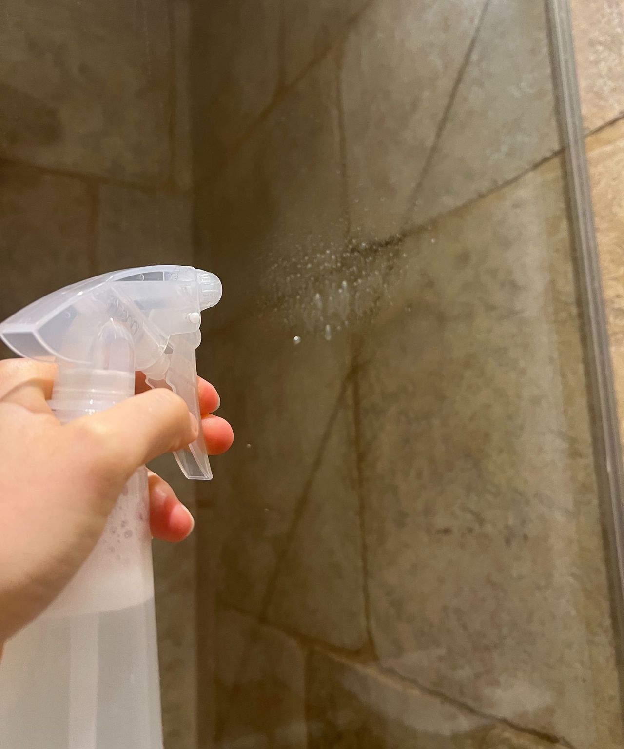 A hand spraying a shower screen with a clear spray bottle with neutral beige tiles behind