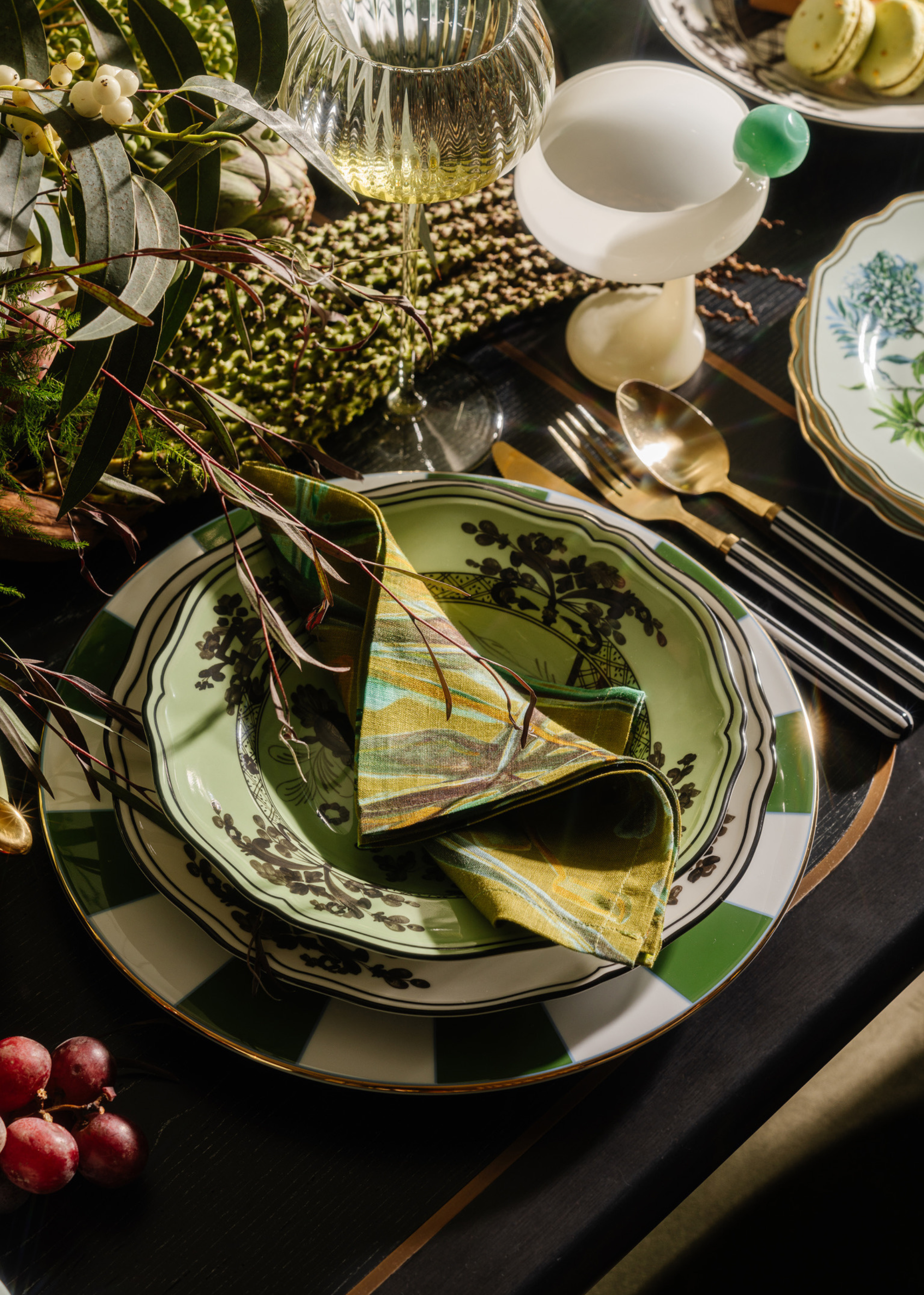 A Christmas table setting with layered decor and rich colors, with multiple plates, a folded napkin, fruit and florals