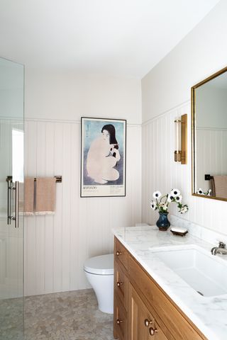 small bathroom with stone floor, wall panneling, marble and timber counter, artwork, glass shower screen and wall light