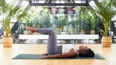 woman lying on an exercise mat on wooden floor sideways to the camera, on her back with legs up at 90 degree angles. there are plants and glass walls behind her.