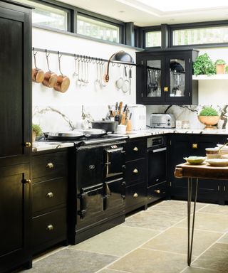 Black and white kitchen with stone flooring, black cabinetry, marble countertops and backsplash, hanging rail with copper pans, plants