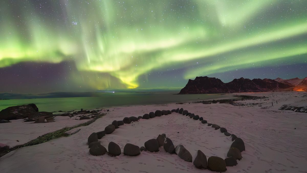 northern lights dance across sky in ribbons of green light while a heart made up of individual stones is laid out on the sandy beach below. 