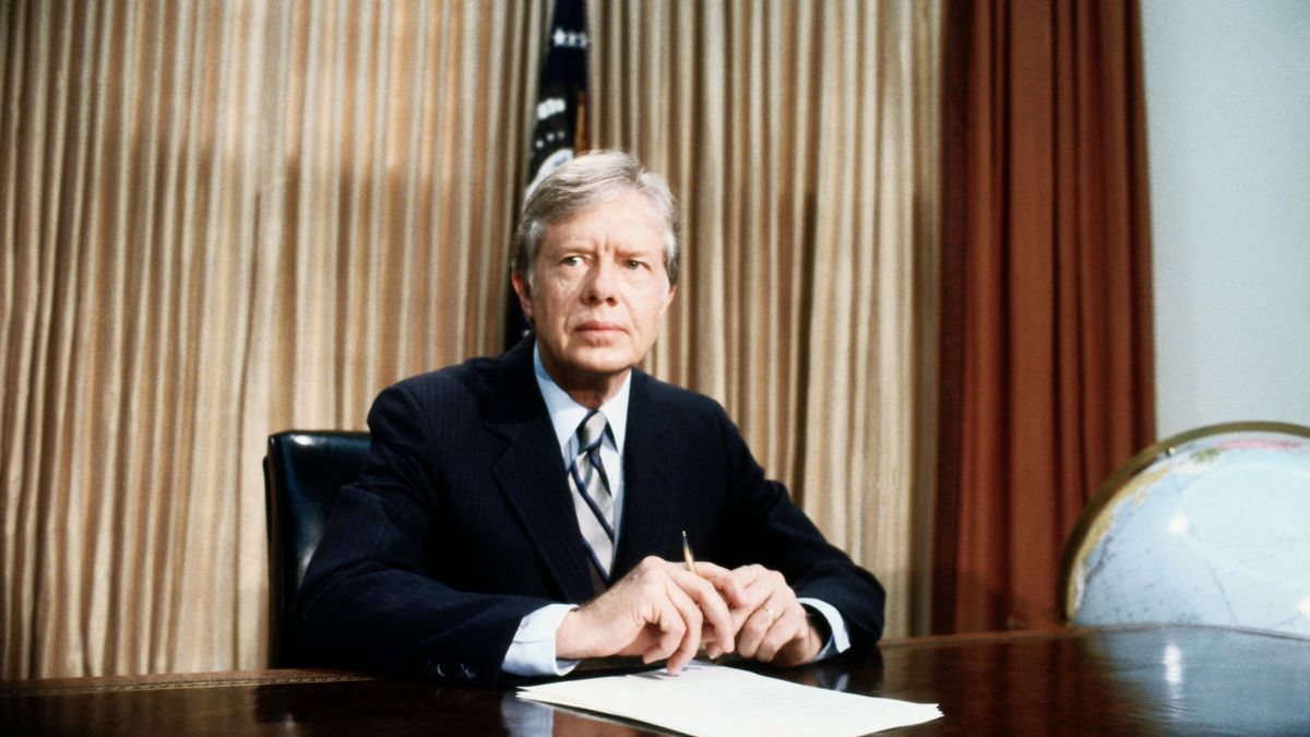 a man in a suit sits at a large wooden desk beside a globe