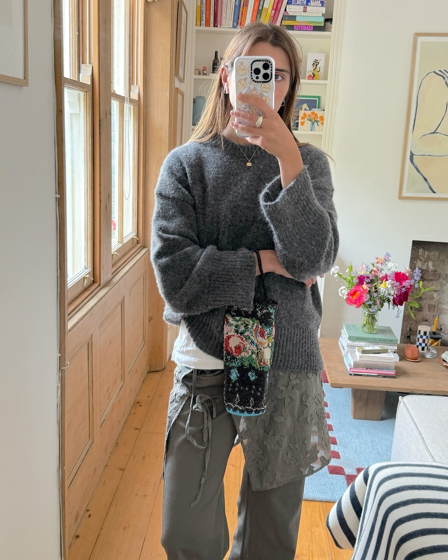 British style influencer Poppy Almond poses for a mirror selfie in her stylish living room wearing a gray sweater, a Cordera beaded floral bag, lace skirt, and pants