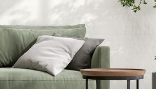 A round wooden side table with black legs next to a sage green sofa with cream and grey cushions on top.