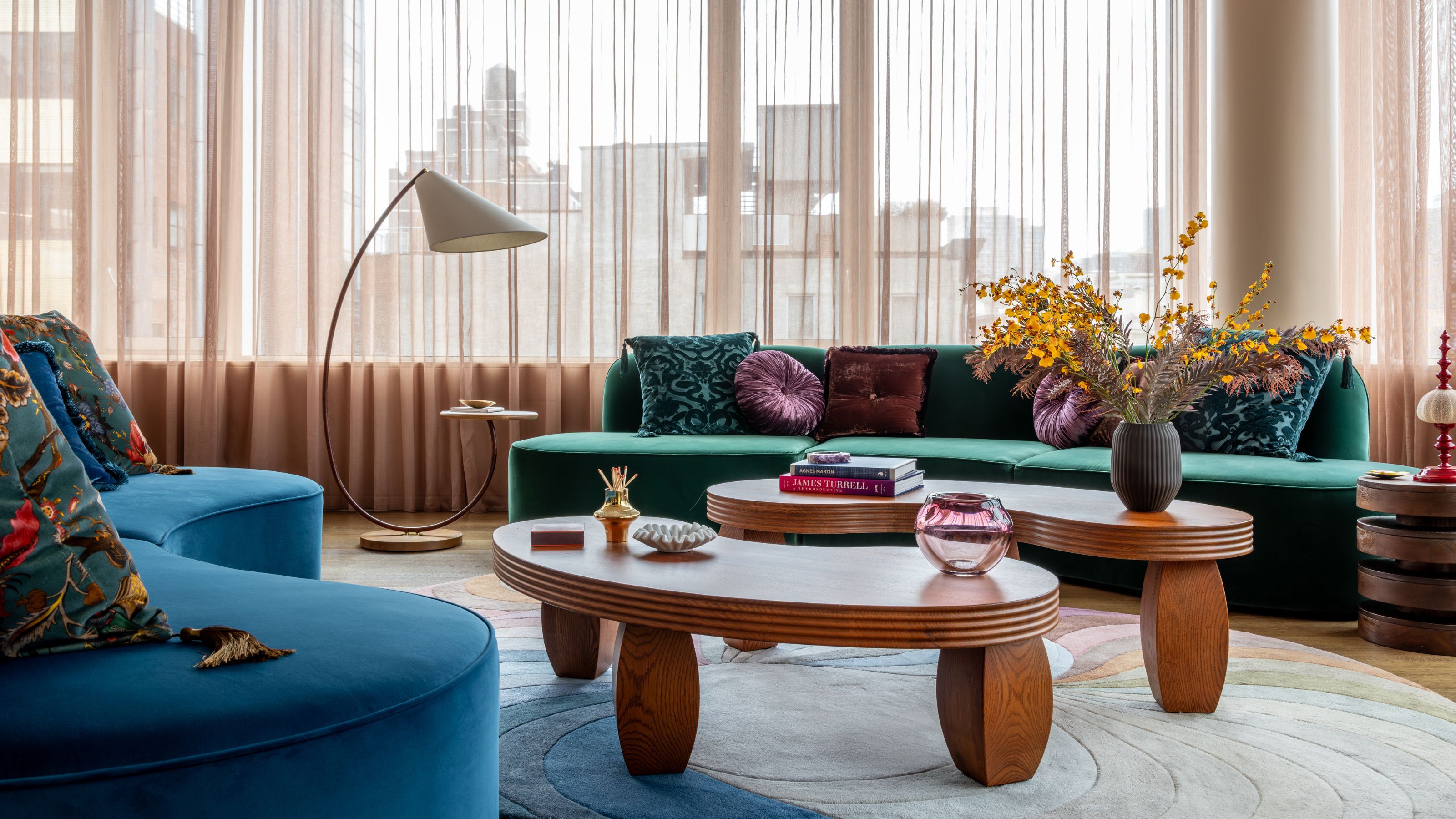 A living room with curving blue sofas, beige curtains, long, oval coffee tables and a round carpet 