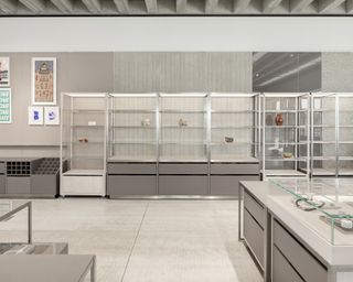 Alternative interior view at the Turner Contemporary shop featuring grey and white walls, light grey flooring, white and grey shelving units and drawers and glass display cases. There are some products on display and framed art on the wall
